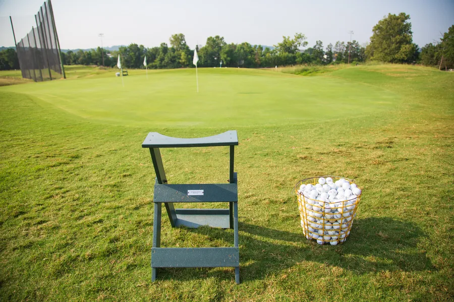 A wire basket of range balls beside a wooden stand, looking out over the practice green and flagsticks at Fairways & Greens