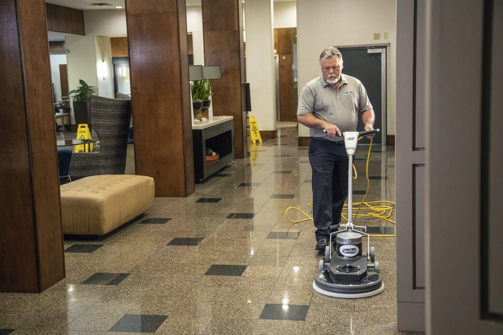 A CleanFreak technician running a floor burnisher across polished granite in a hotel corridor, wet-floor signs set out behind him