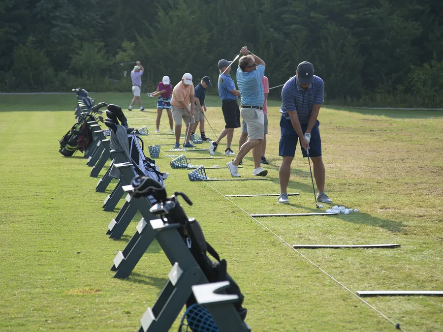 A line of golfers of mixed ages hitting balls from the tee mats at the Fairways & Greens driving range on a bright morning