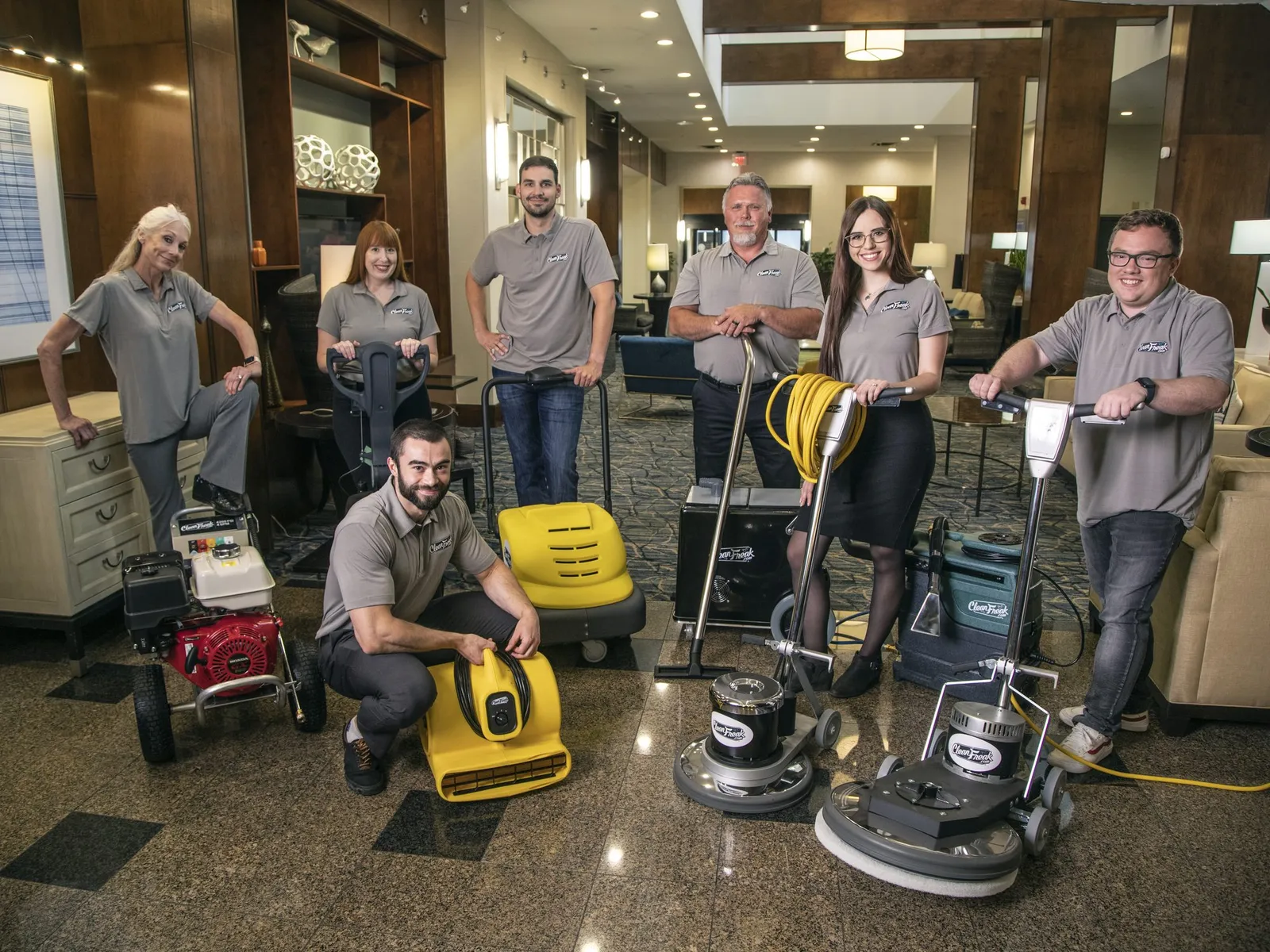 Seven CleanFreak staff in grey branded polos posed around their floor-care range — burnishers, a walk-behind sweeper, an air mover and a pressure washer — in a hotel lobby