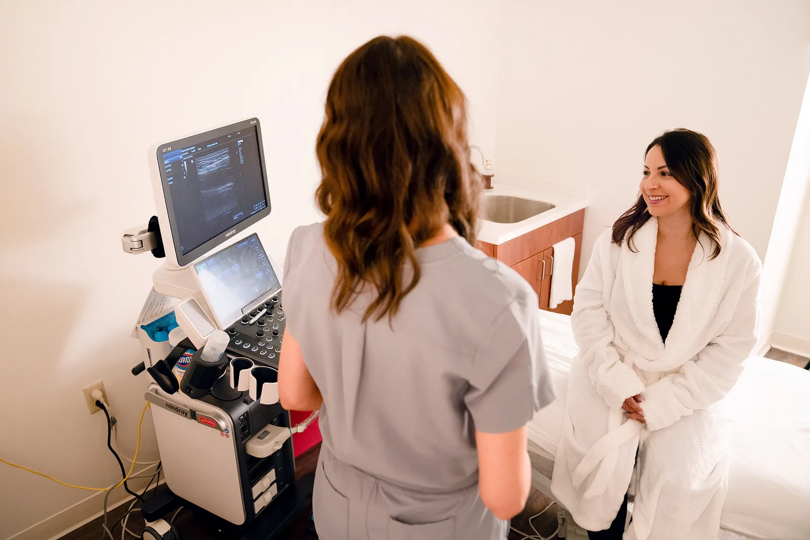 A Mosaic sonographer at the ultrasound console, the scan on screen, with a smiling patient in a robe seated on the exam bed