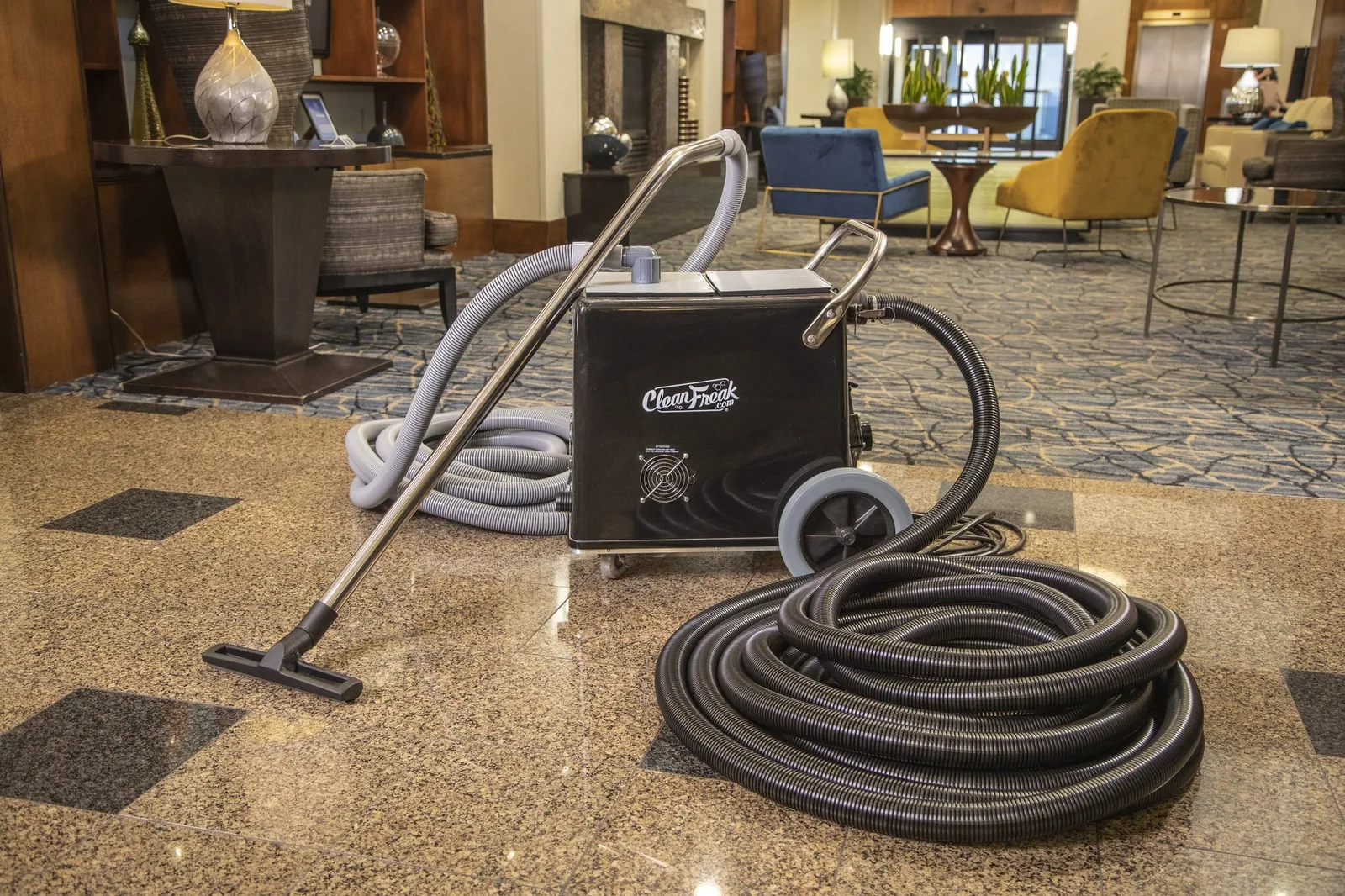 A CleanFreak back-pack style vacuum unit with two coiled hoses standing on a hotel lobby floor